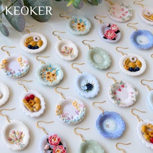 Puede incluir: Una colección de pendientes en miniatura con forma de plato y ganchos dorados. Los diseños incluyen varios motivos como ramos de flores, pasteles variados, bayas y patrones celestiales en colores pastel como azul, rosa y verde.