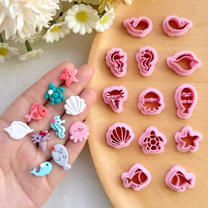 Può includere: Un set di 18 tagliapasta in plastica rosa a forma di varie creature marine, tra cui una stella marina, una conchiglia, un cavalluccio marino, un polpo, una tartaruga e un pesce. I tagliapasta sono esposti su una superficie di legno con una mano che tiene diverse piccole decorazioni di creature marine in pasta di zucchero colorate.