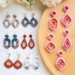 Può includere: Un set di 10 stampi per orecchini in argilla polimerica. Gli stampi sono rosa e presentano vari disegni geometrici e floreali. Gli stampi sono esposti su una superficie di legno con esempi di orecchini realizzati con gli stampi.