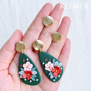 May include: A pair of green teardrop-shaped earrings with floral designs in pink, red, and white. The earrings have gold accents and are displayed on a hand.