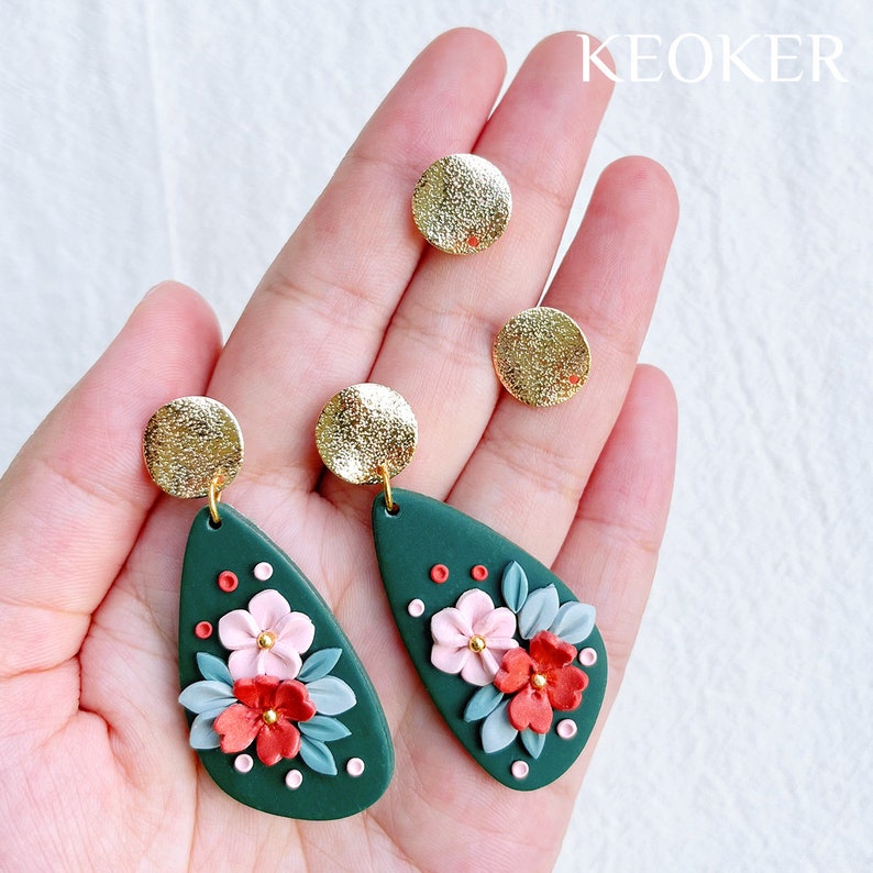 May include: A pair of green teardrop-shaped earrings with floral designs in pink, red, and white. The earrings have gold accents and are displayed on a hand.