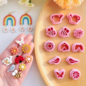 Puede incluir: Un juego de 14 cortadores de galletas de plástico rosa en varias formas, incluyendo flores, mariposas, abejas y regaderas. Los cortadores están dispuestos sobre una superficie de madera. También hay varios artículos decorativos pequeños y coloridos en una mano.