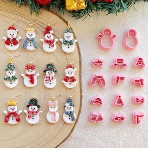 Könnte beinhalten: Eine Sammlung von Schneemann-Ohrringen und Ausstechformen. Die Ohrringe zeigen Schneemänner mit verschiedenen Hüten, Schals und Accessoires. Die Ausstechformen sind rosa und in Form von Schneemännern und Zubehör.