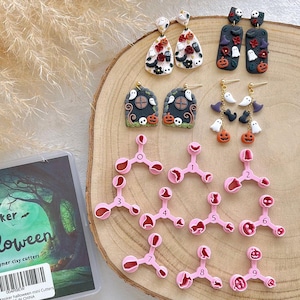 May include: A collection of Halloween-themed polymer clay earrings and cutters. The earrings feature designs of ghosts, pumpkins, and haunted houses. Pink cutters are numbered 1-9. A package with the text "ker halloween" is also present.