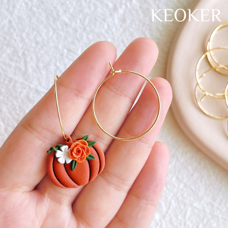May include: A pair of gold hoop earrings with a small orange pumpkin charm. The pumpkin charm has a white flower and a small orange flower on top.