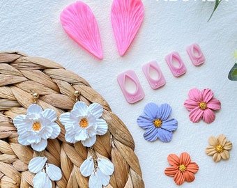 May include: A set of silicone moulds for making polymer clay flowers. The moulds are shaped like flower petals and come in different sizes. The set also includes a set of cutters for making flower centres. The moulds are pink and the cutters are white. The image also shows examples of finished polymer clay flowers.