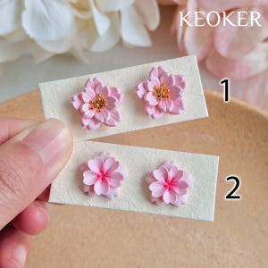 May include: Two pairs of pink flower-shaped stud earrings. The top pair has gold-colored centers, while the bottom pair has bright pink centers. The earrings are displayed on rectangular cards. The word "KEOKER" is printed in white.