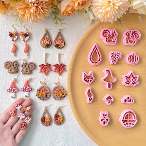 Könnte beinhalten: Eine Sammlung handgefertigter Ohrringe und Ausstecher zum Thema Herbst. Die Ohrringe zeigen Designs wie Füchse, Kürbisse, Eichhörnchen, Pilze und Ahornblätter. Die Ausstecher haben die gleichen Formen. Die Farbpalette umfasst Orange, Braun und Rot.