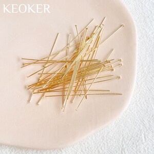 May include: A pile of gold-colored head pins on a light pink surface. The head pins are about 1 inch long.