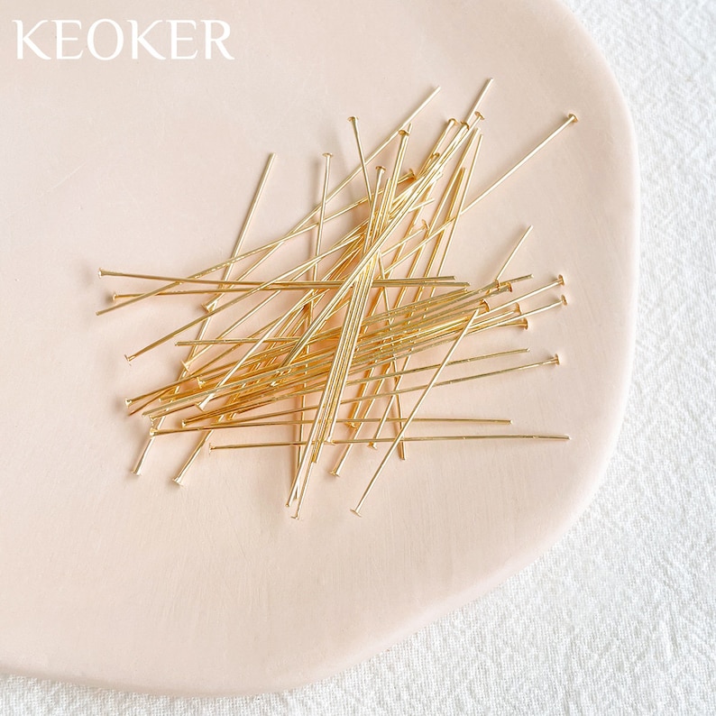 May include: A pile of gold-colored head pins on a light pink surface. The head pins are about 1 inch long.