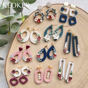 Puede incluir: Una colección de pendientes hechos a mano en varias formas y colores, incluyendo blanco, rosa y azul marino. Muchos pendientes presentan detalles florales y de hojas. Los pendientes se exhiben sobre una superficie de madera. La marca "KEOKER" es visible.