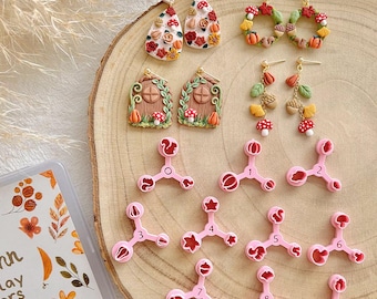 May include: A collection of handmade polymer clay earrings and cutters. The earrings feature autumn-themed designs like pumpkins, mushrooms, and leaves. Pink cutters in various shapes are also visible, along with a clear container with floral designs.