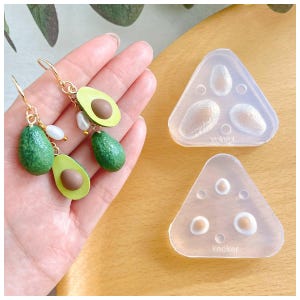 Könnte beinhalten: Ohrringe im Avocado-Design mit goldfarbenen Haken und weißen Perlenakzenten. Die Ohrringe zeigen grüne Avocadohälften und ganze Avocados. Zwei dreieckige Silikonformen sind ebenfalls sichtbar, die zur Herstellung von avocadoförmigen Gegenständen bestimmt sind.