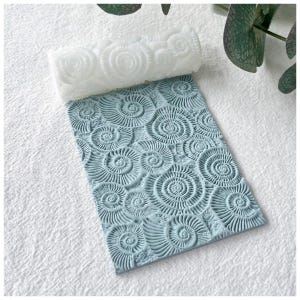 May include: A white rolling pin and a light blue rectangular mat, both featuring a spiral seashell pattern. The rolling pin is cylindrical, and the mat is covered in a repeating design of the same spiral motif. The items are on a white surface.