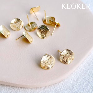 May include: Gold-toned metal earring blanks with a textured finish. The blanks are round and have a small hole in the centre. The blanks are arranged on a light pink surface. KEOKER is printed in the top right corner of the image.