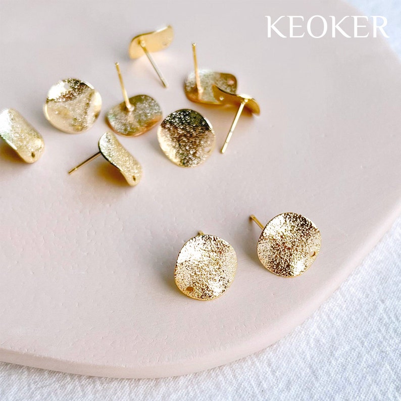 May include: Gold-toned metal earring blanks with a textured finish. The blanks are round and have a small hole in the center. The blanks are arranged on a light pink surface. KEOKER is printed in the top right corner of the image.