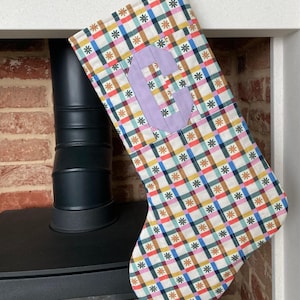 May include: A Christmas stocking featuring a colourful plaid design in red, blue, yellow, and green. A lavender letter "G" is sewn onto the front. The stocking has a loop for hanging.