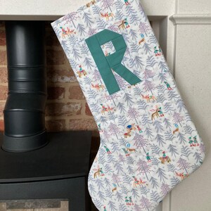 May include: A festive Christmas stocking with a white background, patterned with deer, trees, and small figures. A teal letter "R" is appliquéd on the front. The stocking is hanging near a fireplace.