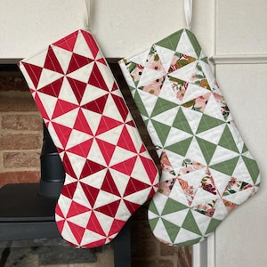 Könnte beinhalten: Zwei Weihnachtsstrümpfe hängen an goldenen Haken. Einer hat ein rot-weißes geometrisches Muster, der andere ein grün-blumiges Design. Beide sind gesteppt und haben einen weißen Rand.