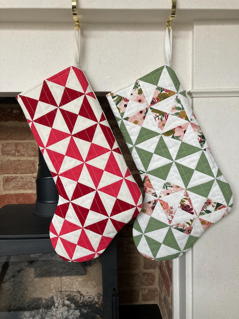 以下が含まれることがあります： 金色のフックに吊るされた2つのクリスマスストッキング。1つは赤と白の幾何学模様、もう1つは緑と花の模様です。どちらもキルティングで、白いトリムが付いています。お祝いの家の装飾。