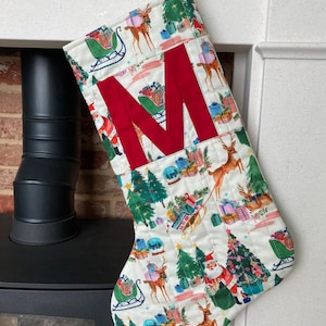 May include: Christmas stocking with a festive pattern featuring reindeer, Santa, and gifts on a white background. A large red letter "M" is sewn on the front. The stocking has a green and white patterned loop for hanging.