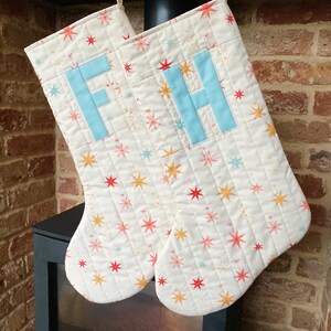 May include: Two cream-coloured quilted Christmas stockings, patterned with colourful stars. Each stocking features a light blue letter, one with an "F" and the other with an "H".