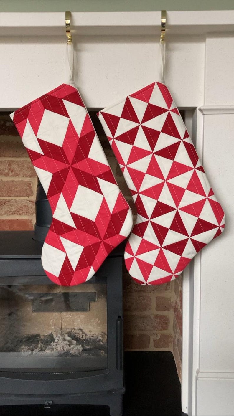 以下が含まれることがあります： 赤と白の幾何学模様のクリスマスストッキングが2つ、暖炉に吊るされています。1つはスターバーストのデザイン、もう1つは三角形のパターンです。ストッキングはキルト生地で作られています。