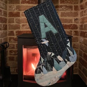 May include: A navy blue Christmas stocking with a teal letter "A" appliqué. The stocking features a winter forest scene with owls, birds, and rabbits. The background has a starry night design. The stocking is hanging near a fireplace.