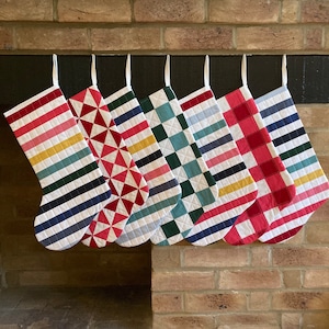 May include: Seven colourful Christmas stockings hanging on a fireplace mantel. The stockings are made of fabric and have different patterns, including stripes, polka dots, and geometric designs. The stockings are in shades of red, blue, green, yellow, and white.