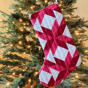 May include: A red and white quilted Christmas stocking with a geometric pattern. The stocking is hanging on a Christmas tree.