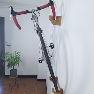 Könnte beinhalten: Ein Rennrad mit rotem Lenkerband ist vertikal an einer weißen Wand mit zwei hellbraunen Wandhalterungen aus Holz befestigt. Das Fahrrad verfügt über schwarze Reifen, silberne Metallkomponenten und schwarze Pedale, die sicher von den Halterungen gehalten werden.