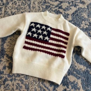 May include: A white sweater featuring the American flag. The flag design includes a navy blue rectangle with white stars and alternating burgundy and white stripes. The sweater has a crew neck and long sleeves.