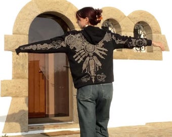 May include: A black hooded sweatshirt with a gray graphic design on the back. The design appears to be a stylized emblem or symbol. The person is wearing blue jeans.