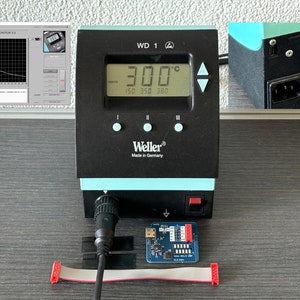 May include: A black and teal Weller WD 1 soldering station with a digital display showing 300°C. The display also shows temperature presets. A small circuit board and ribbon cable are in front of the station. Made in Germany.
