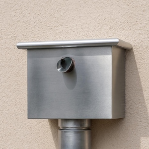 May include: A rectangular, brushed stainless steel rain gutter collector box featuring a smooth, flat top edge and a small, circular side outlet. The metal box is mounted vertically and connects to a cylindrical downspout pipe below it.