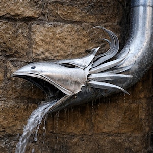Op de afbeelding: Een zilverkleurige metalen regenpijpverlenging in de vorm van een vogel- of drakenkop met een open snavel, waaruit water naar beneden stroomt. Het metalen beeldhouwwerk heeft puntige, veerachtige vinnen aan de zijkant en een gebogen nek.