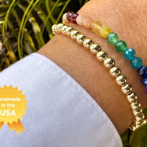May include: Two gold bracelets with colorful gemstone beads. The top bracelet features red, orange, yellow, green, blue, and purple beads. The bottom bracelet is a simple gold chain.