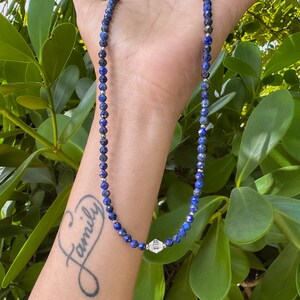 May include: A blue gemstone necklace with a silver charm. The necklace is on a person's wrist, and the charm is in the center of the image. The person has a tattoo that says "Family" on their wrist.