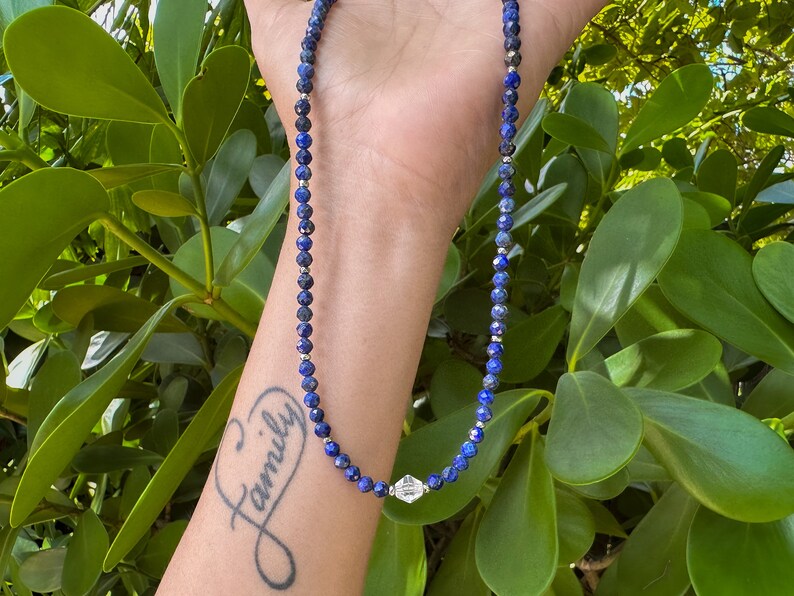 May include: A blue gemstone necklace with a silver charm. The necklace is on a person's wrist, and the charm is in the center of the image. The person has a tattoo that says "Family" on their wrist.