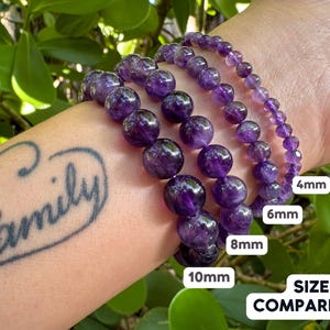 May include: Four amethyst bracelets of different sizes, 4mm, 6mm, 8mm, and 10mm, are shown on a person's wrist. The bracelets are stacked on top of each other, with the smallest bracelet on top and the largest bracelet on the bottom. The text "SIZE COMPARISON" is visible below the bracelets.