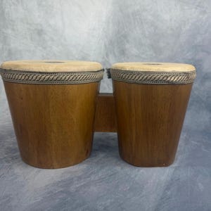 May include: A set of two bongo drums connected by a central wooden bridge. Each drum features a smooth, light brown wooden shell and a light-colored, circular drumhead secured with a decorative, braided fabric trim around the top edge.