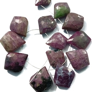 May include: A strand of faceted, irregular-shaped gemstones in shades of deep red and purple. Some stones have green and black inclusions. The gemstones are strung on a thin, silver-colored wire, and are displayed against a white background.