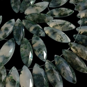 May include: Close-up of moss agate beads, marquise-shaped and strung on a white cord. The translucent stones display green and white patterns. The beads are suitable for jewellery making, set against a black backdrop.