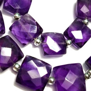 Op de afbeelding: Close-up van een ketting met vierkante, gefacetteerde amethist kralen. De dieppaarse edelstenen zijn geregen met kleine zilverkleurige kralen. De ketting wordt gepresenteerd tegen een witte achtergrond, waardoor de kleur en vorm van de kralen worden benadrukt.