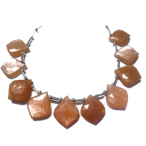 May include: A strand of faceted, peach-colored gemstone beads. The beads are diamond-shaped and strung together with silver-colored metal beads and wire. The gemstones have a translucent quality and reflect light.