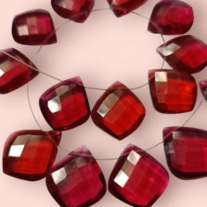May include: A strand of faceted, deep red, cushion-shaped beads arranged in a circular pattern. Each translucent bead features multiple geometric facets that reflect light, creating a sparkling appearance. The beads are strung on a thin, metallic wire.