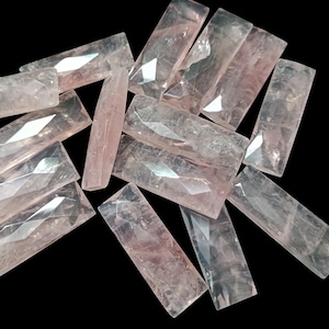 May include: A group of 14 faceted, translucent, light pink, rectangular gemstone beads. The beads are arranged in a random pile.
