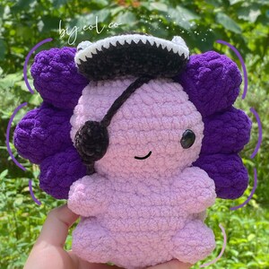 May include: A purple and pink crocheted axolotl plush toy wearing a black and white cowboy hat with a black eye patch. The toy is sitting on a hand.