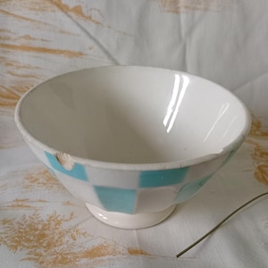 May include: White ceramic bowl with a light blue chequered pattern around the rim. The bowl has a small foot and a chip on the edge. The bowl is on a white tablecloth with a yellow floral design.