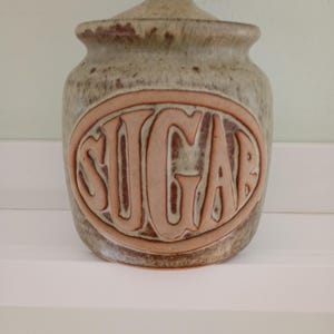 May include: A ceramic sugar jar with a lid. The jar is a speckled beige colour with a brown rim and a light brown oval with the word "SUGAR" in raised letters. The jar is sitting on a white shelf.